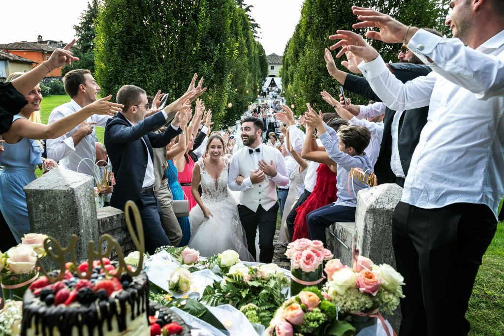 Bride and groom joyfully walk through a cheering crowd of guests at an outdoor wedding celebration, surrounded by flowers and a decorated cake.