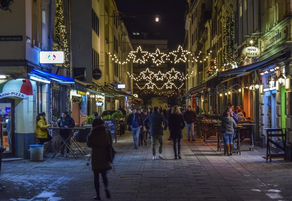 People walking along a lively, well-lit street at night with festive star-shaped lights overhead and outdoor diners enjoying food and drinks at cafes and restaurants.