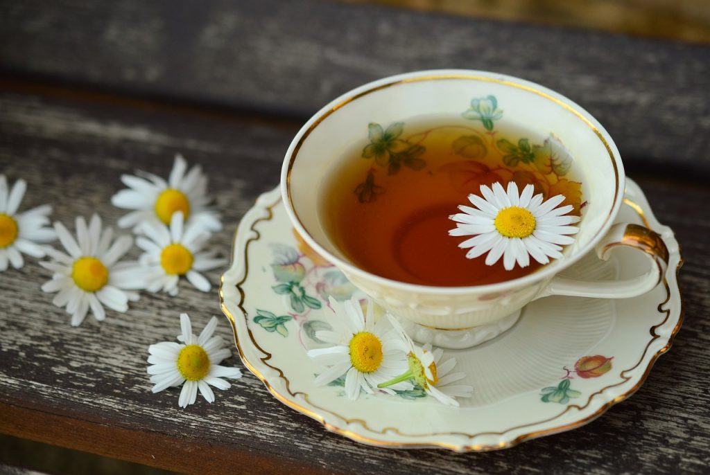 A delicate floral china teacup filled with chamomile tea, garnished with a fresh chamomile flower, resting on a matching saucer with more chamomile flowers scattered on a rustic wooden surface.