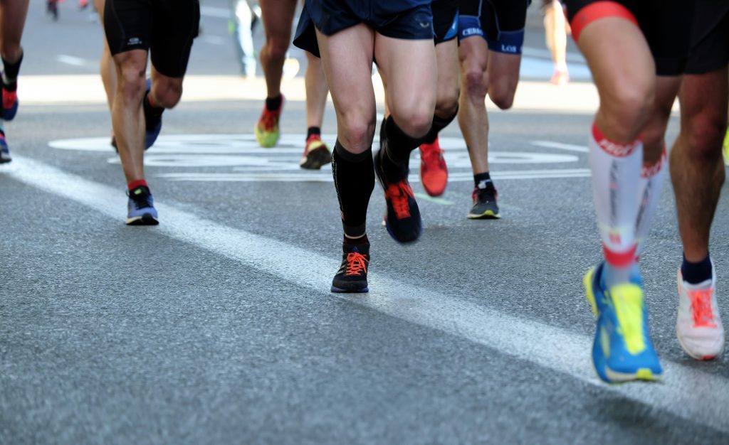 A group of runners mid-stride during a road race, with a close-up focus on their legs and running shoes as they move along a marked asphalt street.