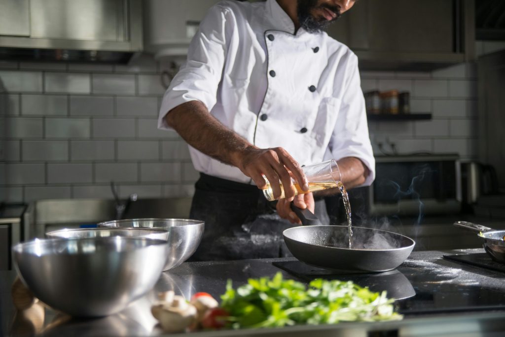 Chef in a commercial kitchen pouring oil into a frying pan, preparing for cooking, with metal bowls and fresh herbs on the counter.