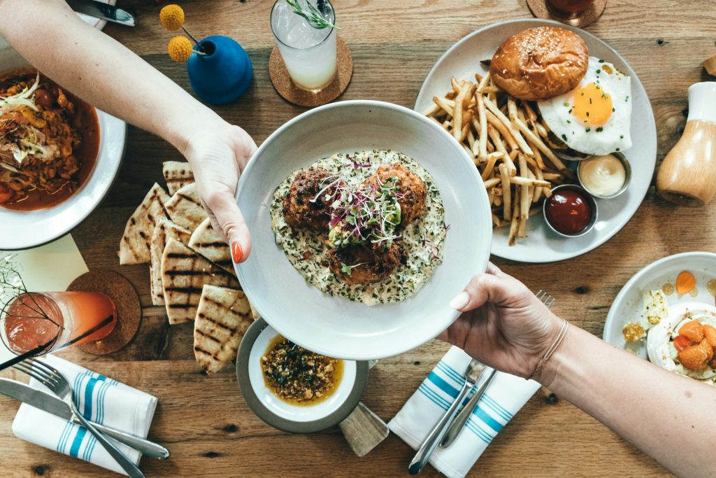 Overhead view of a shared family-style meal at a restaurant, featuring a variety of vibrant dishes including fries, a burger with a fried egg, grilled flatbreads, a creamy main dish being passed between hands, and colourful drinks on a wooden table.