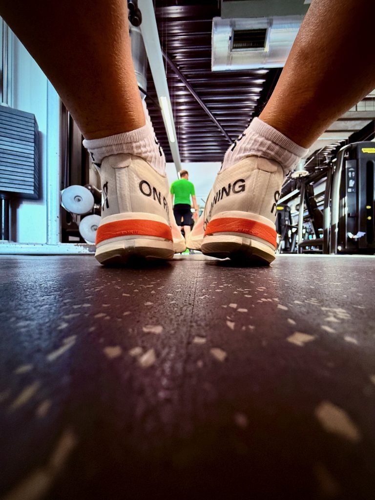Close-up of white On Running trainers resting on the gym floor, with a person walking in the background and gym equipment visible