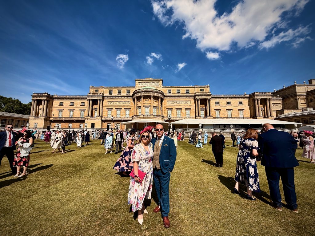 Christopher Blackburn and his wife standing together on the lawn at the Buckingham Palace Garden Party, dressed in formal attire among a crowd of guests. The palace's grand rear facade and a bright blue sky with scattered clouds are visible in the background.