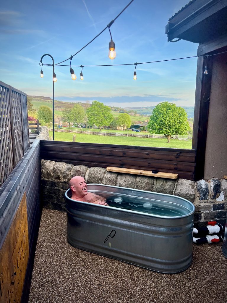 A man smiling while immersed in a cold plunge tub outdoors at The Paddock Wellness Club, with panoramic views of the Calder Valley and festoon lights overhead.