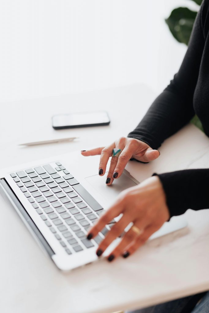 Close-up of a person using a laptop on a white desk, wearing dark nail polish and a turquoise ring, with a smartphone and pen placed nearby.