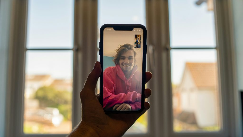 A person holding a smartphone during a video call with a smiling young man wearing a red hoodie, with bright natural light coming through large windows in the background.