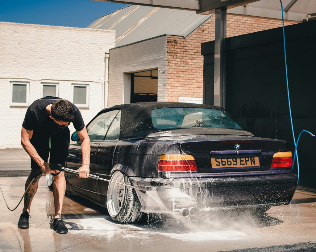 A man wearing a black t-shirt and shorts is using a pressure washer to clean the rear side of a black BMW convertible covered in soap, at an outdoor car wash station on a sunny day.