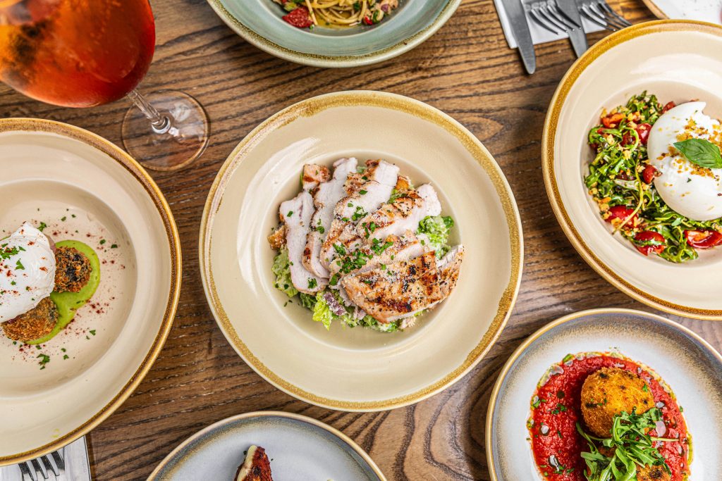 A rustic wooden table set with an elegant spread of dishes, featuring a central plate of sliced grilled chicken atop a Caesar-style salad with romaine lettuce and grated cheese. Surrounding plates include burrata with tomato and rocket salad, crispy arancini in tomato sauce, poached egg with falafel and pea purée, and a pasta dish with cherry tomatoes. A glass of rosé adds a refreshing touch to the vibrant, restaurant-style presentation.