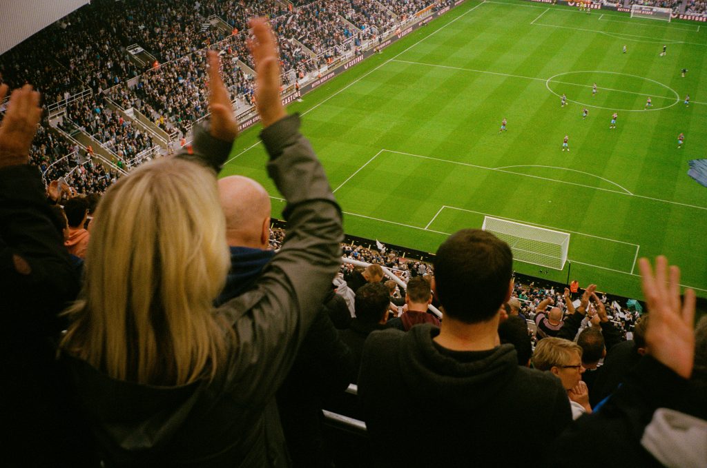 Premier League football fans cheering in a packed stadium during a live match, viewed from the stands behind the goal.