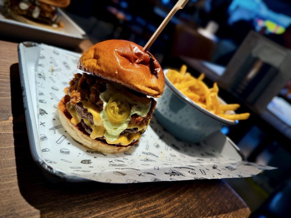 Close-up of the Born Slippy burger at Fat Hippo Leeds, featuring double patties, melted American cheese, braised pork rib meat, jalapeños, garlic butter mayo, and a toasted brioche bun, served with a side of seasoned fries in a blue enamel bowl.