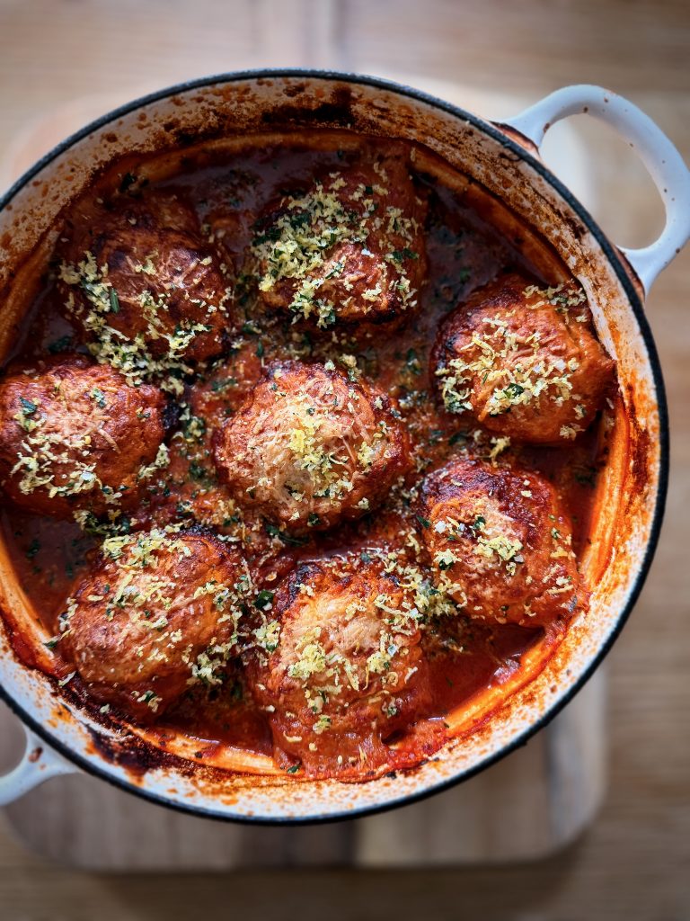 Golden parmesan dumplings baked in a rich tomato arrabbiata stew, served in a white enamel casserole dish with a bubbling, caramelised edge and garnished with lemon zest and fresh herbs.