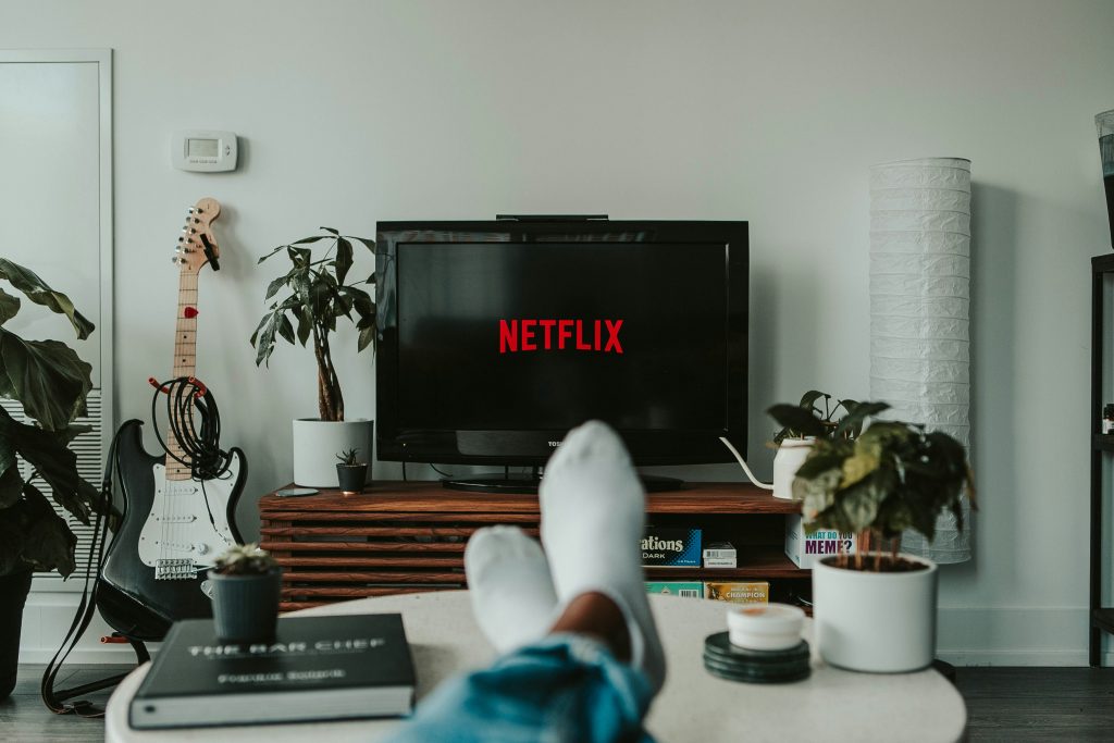 Person relaxing at home with feet up, watching Netflix on a flat-screen TV in a modern living room with plants, a guitar, and board games in view.
