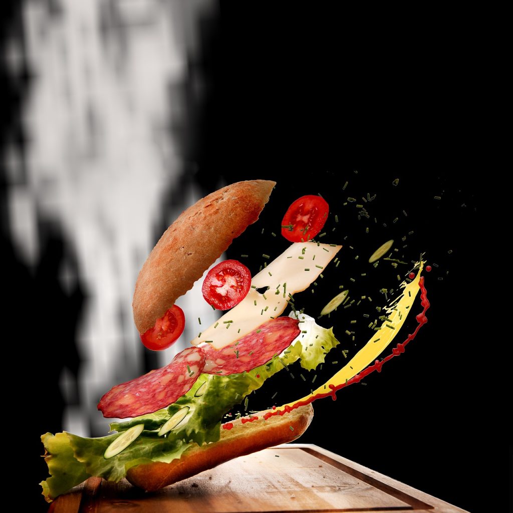 Deconstructed sandwich with ingredients floating mid-air, including a baguette, lettuce, cucumber, salami, cheese, tomato slices, herbs, mustard, and ketchup, against a dramatic black background with motion blur effects.