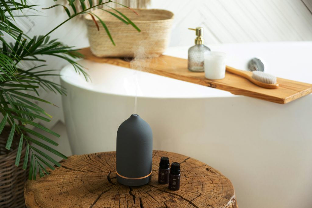 A black ceramic essential oil diffuser emitting mist, placed on a rustic wooden stool beside a white freestanding bathtub. Nearby are two small essential oil bottles, a wooden bath brush, a candle, and a glass soap dispenser on a wooden bath tray. Lush green plants and a woven basket complete the calming spa-like bathroom setting.