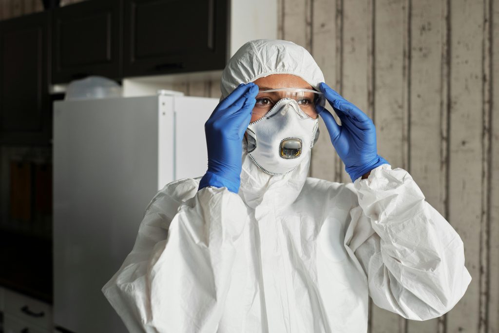 Person wearing full protective gear, including a white hazmat suit, gloves, safety goggles, and a respirator mask, preparing for an asbestos survey in a residential setting.