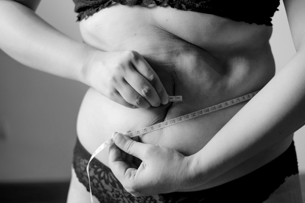 Black and white photo of a woman in underwear measuring her waist with a tape measure, highlighting body image, weight loss, and health awareness themes.