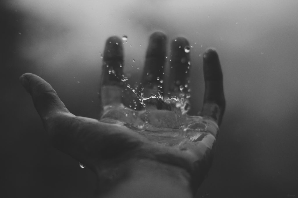 A black and white close-up photo of an open hand catching raindrops, with water splashing on the palm against a blurred background.