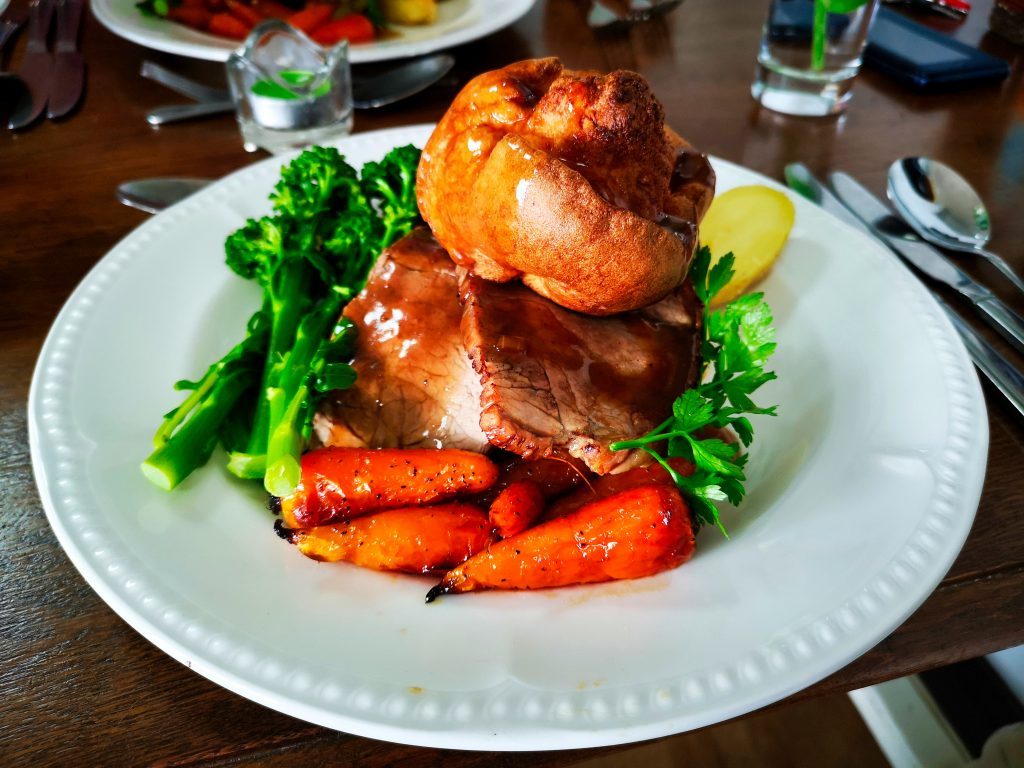 A traditional British roast dinner served on a white plate, featuring slices of roast beef topped with a golden Yorkshire pudding, glazed carrots, tenderstem broccoli, parsley garnish, and a slice of boiled potato.