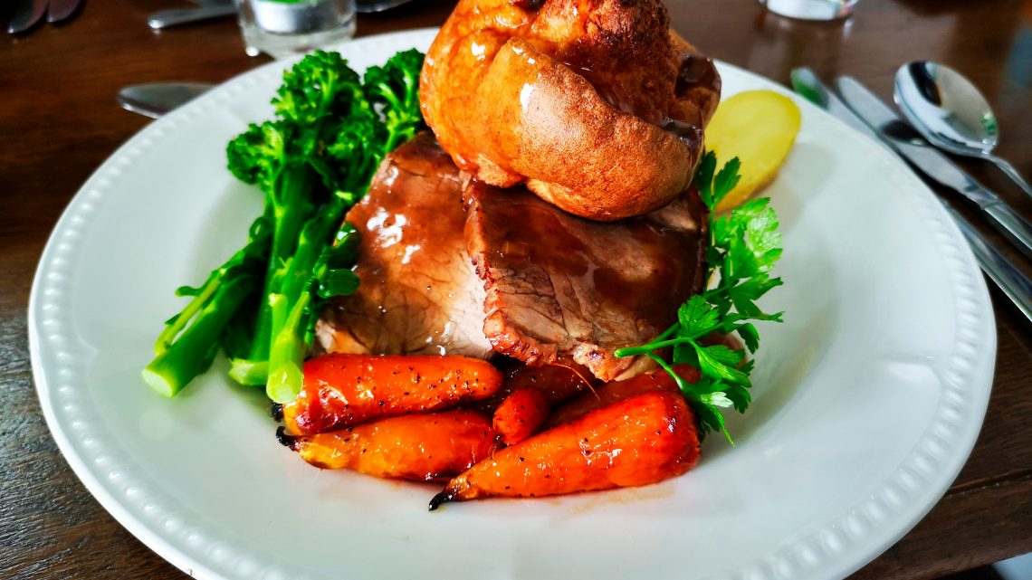 A traditional British roast dinner served on a white plate, featuring slices of roast beef topped with a golden Yorkshire pudding, glazed carrots, tenderstem broccoli, parsley garnish, and a slice of boiled potato.