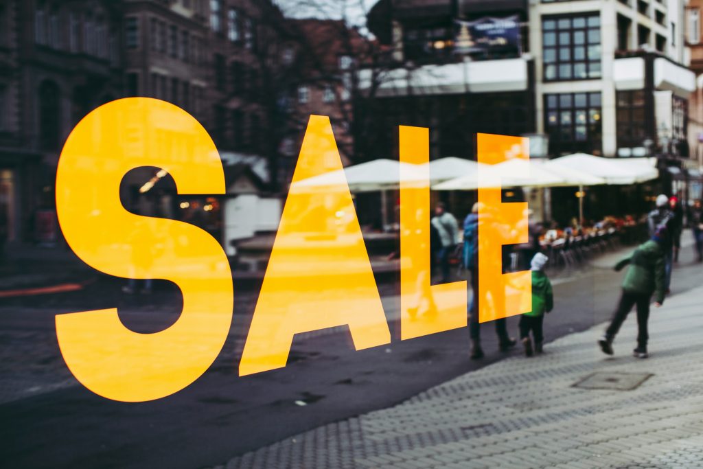 Large yellow “SALE” text on a shop window, reflecting a busy pedestrian street with people walking and outdoor seating visible.