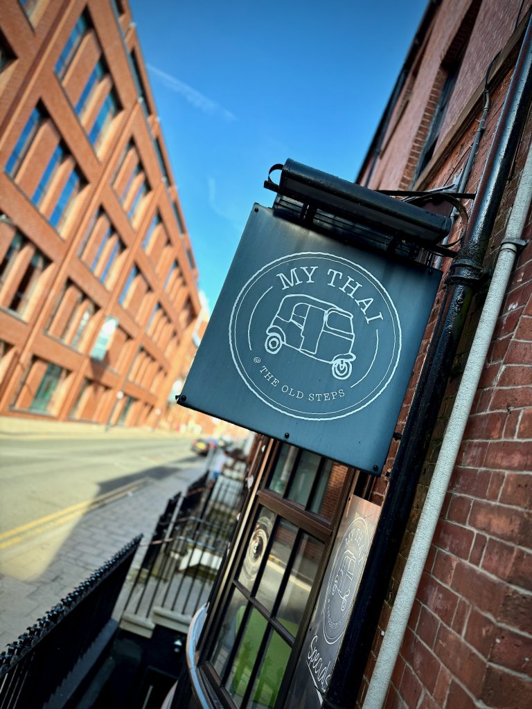 Sign for My Thai at The Old Steps in Leeds, mounted on a red brick building along York Place, with city street in the background.