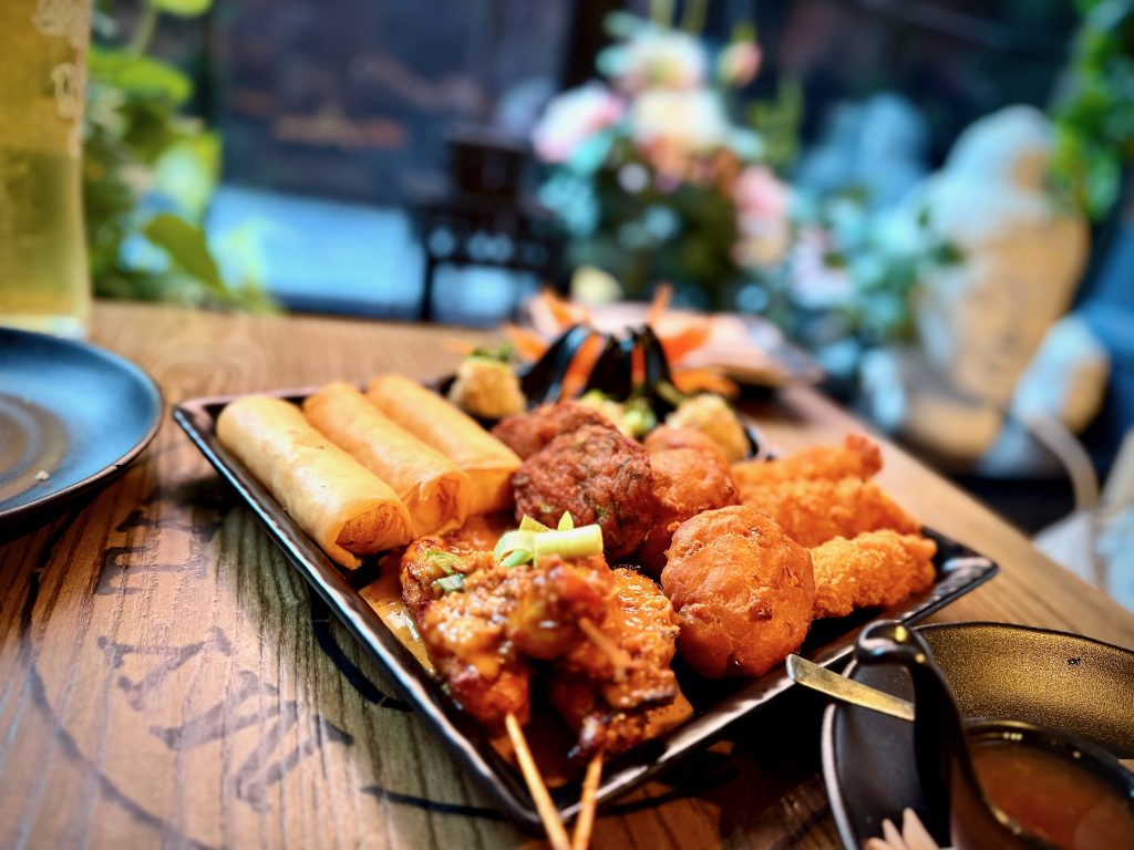 Close-up of My Thai’s sample platter featuring spring rolls, chicken satay, fish cakes, corn cakes, and dumplings, served on a wooden table with floral decor in the background.