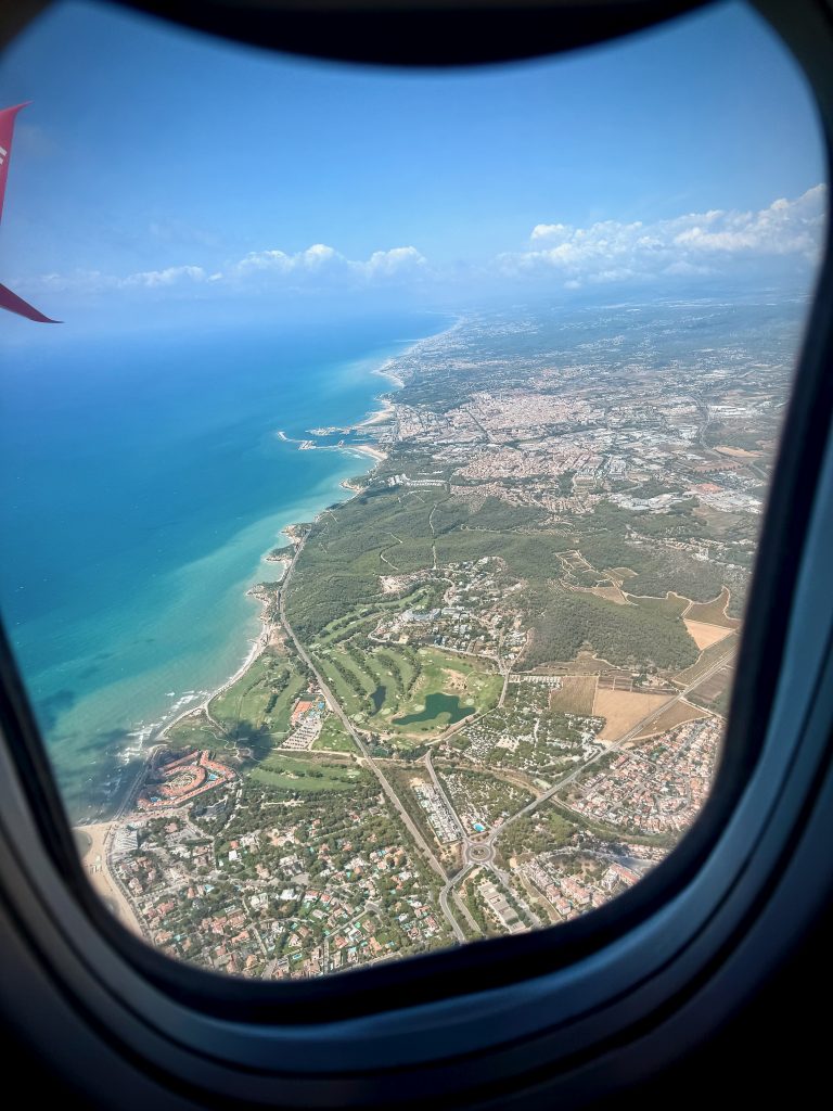 Aerial view of the Spanish coastline and cityscape near Barcelona, photographed through an airplane window with the turquoise Mediterranean Sea stretching alongside the land.