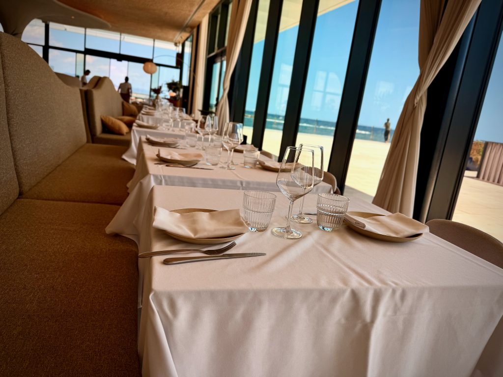 Elegant seaside restaurant interior with neatly set tables covered in white cloths, wine glasses, and folded napkins, overlooking large windows with a view of the ocean outside.