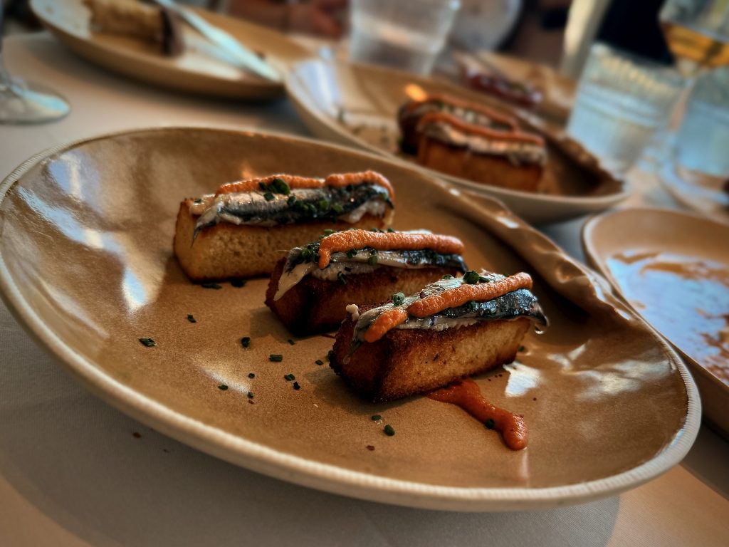 Close-up of a gourmet dish featuring toasted bread topped with anchovies, a drizzle of red pepper sauce, and garnished with fresh herbs, served on a beige ceramic plate in a fine dining restaurant.