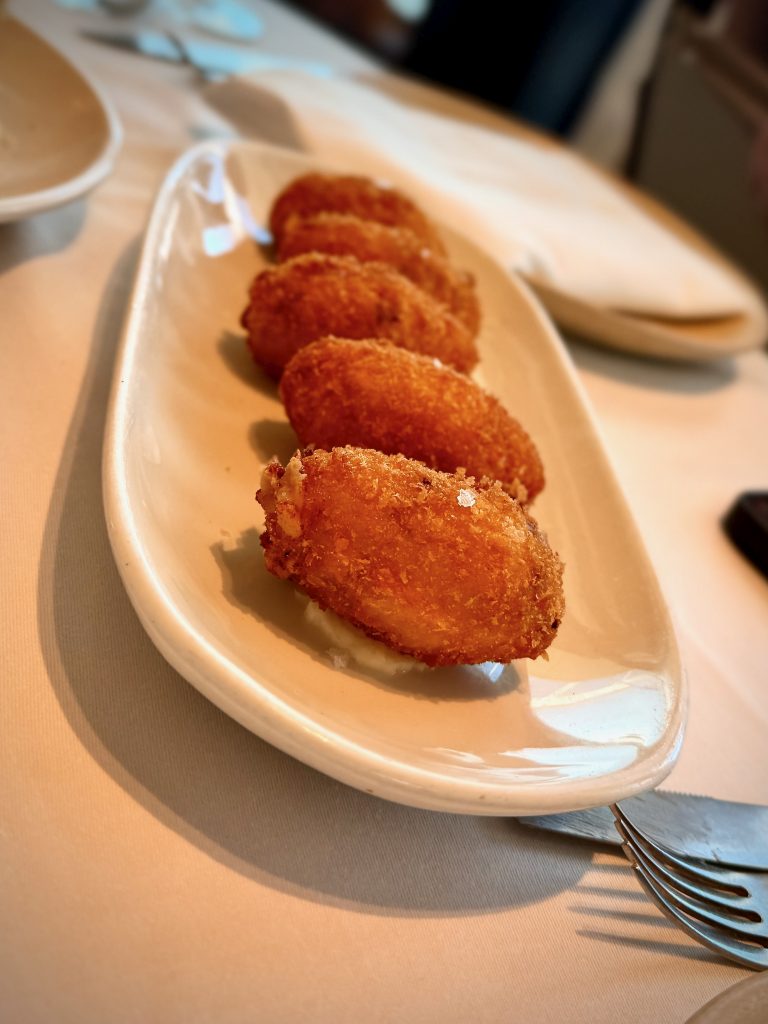 Close-up of a ceramic plate with golden, crispy Spanish croquettes arranged in a row, served on a white tablecloth in a fine dining restaurant.