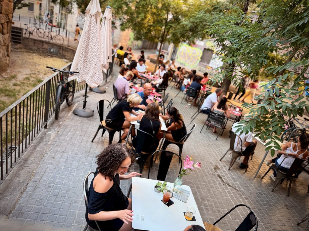 Outdoor terrace dining in Barcelona, with groups of people enjoying food and drinks at tables shaded by trees and parasols on a lively street.