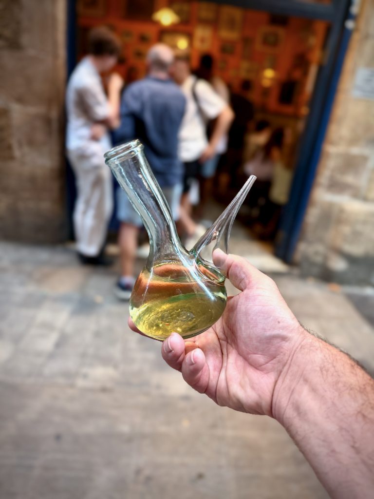 Hand holding a traditional Catalan porró wine pitcher filled with white wine, with blurred background of people socialising outside a rustic bar in Barcelona.