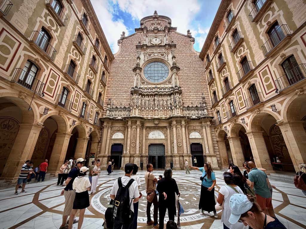 Tourists gathered in the ornate courtyard of Montserrat Monastery in Catalonia, Spain, admiring the grand basilica façade with intricate stone carvings and a large circular stained glass window.