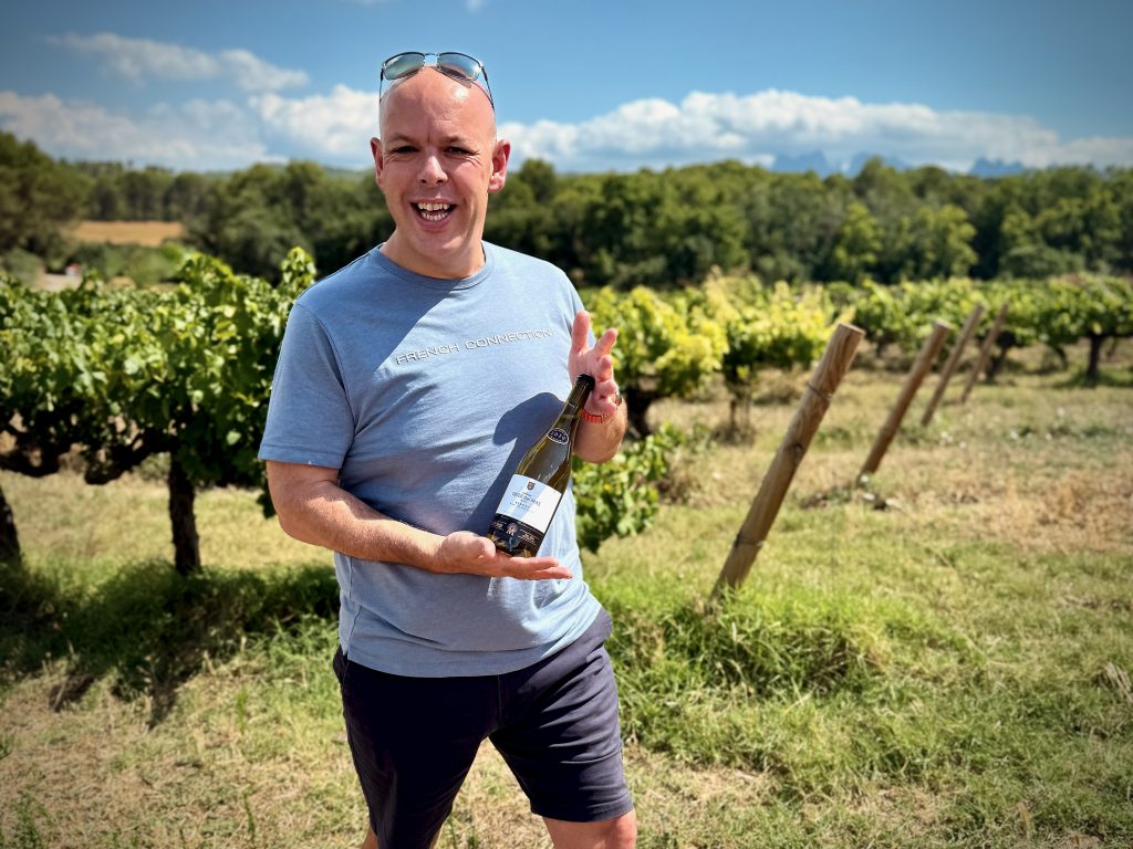Smiling man standing in a vineyard on a sunny day, holding a bottle of wine and wearing a light blue French Connection t-shirt and sunglasses on his head.