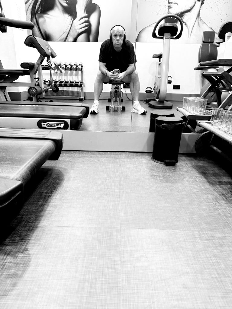 Black and white photo of a man sitting on a bench in a gym, wearing headphones and holding a phone, with dumbbells, treadmills, and fitness equipment visible around him.