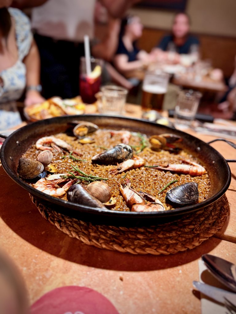 Close-up of a traditional Spanish seafood paella served in a large pan, topped with mussels, clams, and prawns, on a restaurant table with drinks and people dining in the background.
