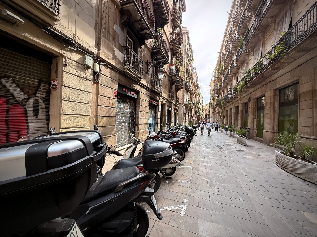 Narrow street in Barcelona lined with scooters and motorbikes, surrounded by historic buildings with balconies and plants, and people walking in the distance.