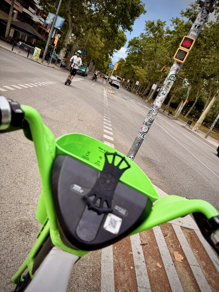 View from a green rental bike at a city cycle lane in Barcelona, with scooters and cyclists using the road, trees lining the street, and traffic lights ahead.