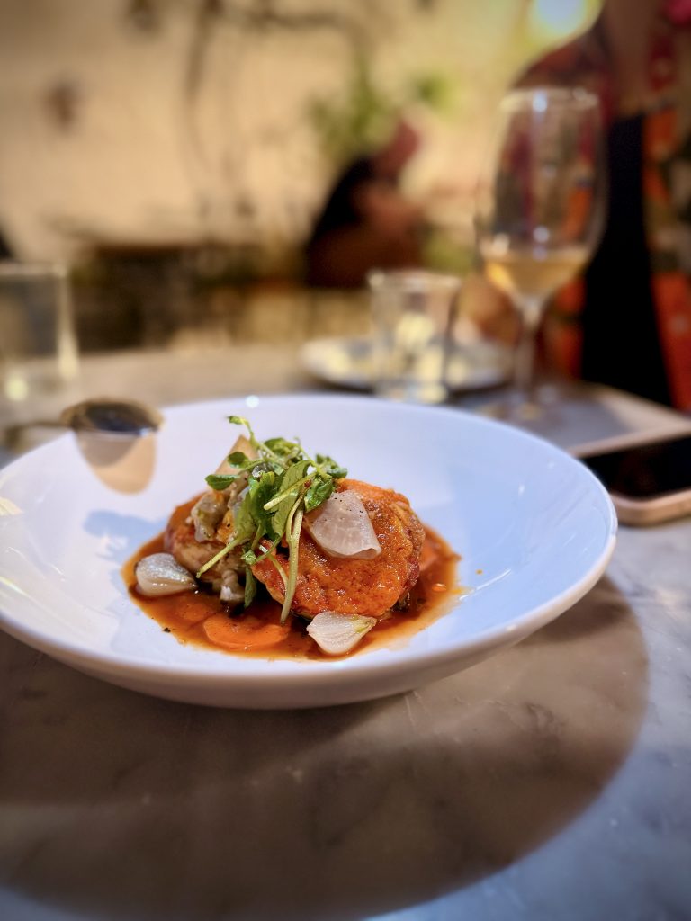 A gourmet dish served on a white plate, featuring a golden-brown patty topped with microgreens and thinly sliced vegetables, presented in a rich red sauce, with a wine glass and blurred restaurant background.