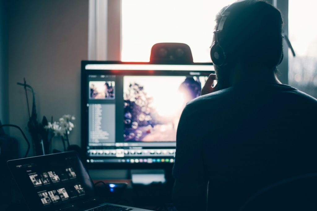 A person wearing headphones sits at a desk, working on a computer with a large monitor displaying an image editing program. A laptop with a dark interface is open in the foreground, and soft daylight streams through a window behind the setup.