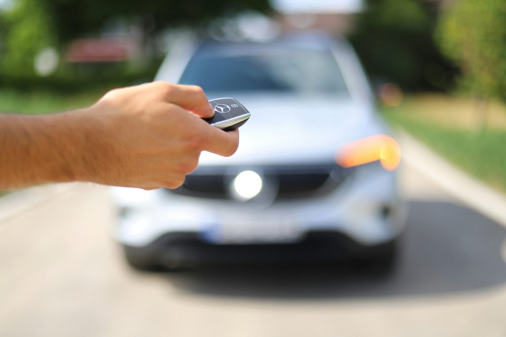 Close-up of a hand holding a Mercedes-Benz car key fob, with a blurred white Mercedes vehicle in the background and its indicator light illuminated.