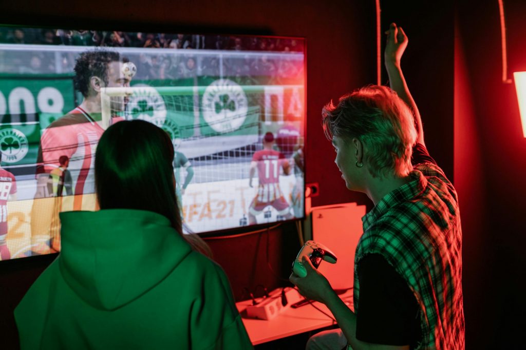 Two people playing a football video game on a large screen, one holding a controller and raising an arm in celebration.