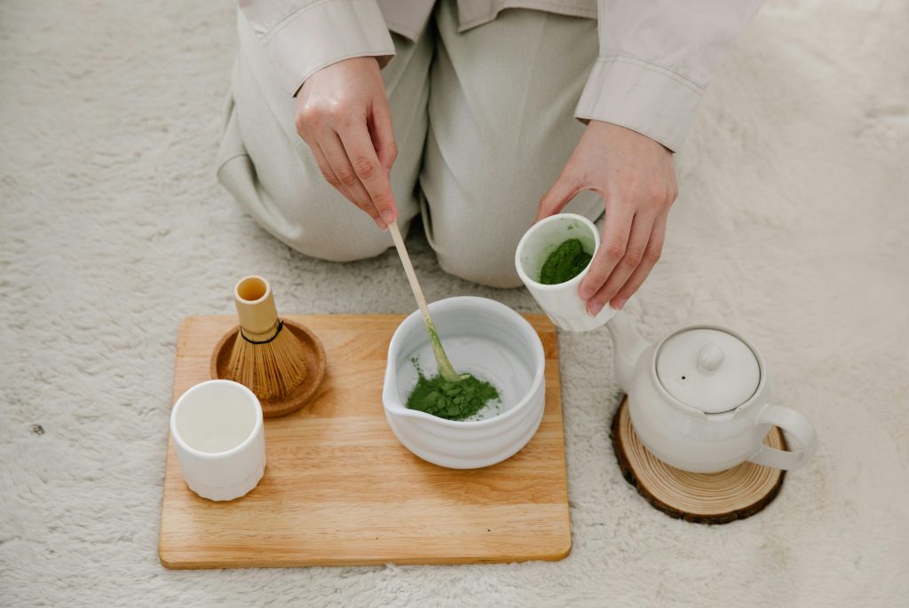 Person preparing green matcha or kratom powder in a white bowl with a bamboo whisk and teapot on a wooden tray.