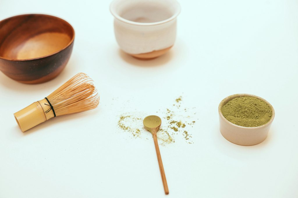 Flat lay of green powder with a bamboo whisk, wooden spoon, and ceramic bowls on a light background.
