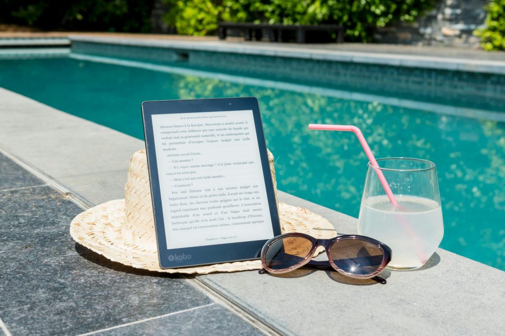 An e-reader on top of a straw hat beside sunglasses and a glass of lemonade with a pink straw, set next to a swimming pool.