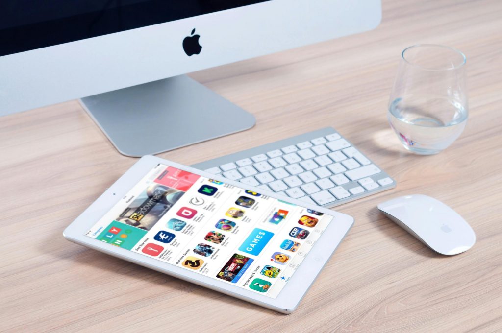 Tablet displaying gaming and app store on a wooden desk with an Apple iMac, keyboard, mouse, and a glass of water.