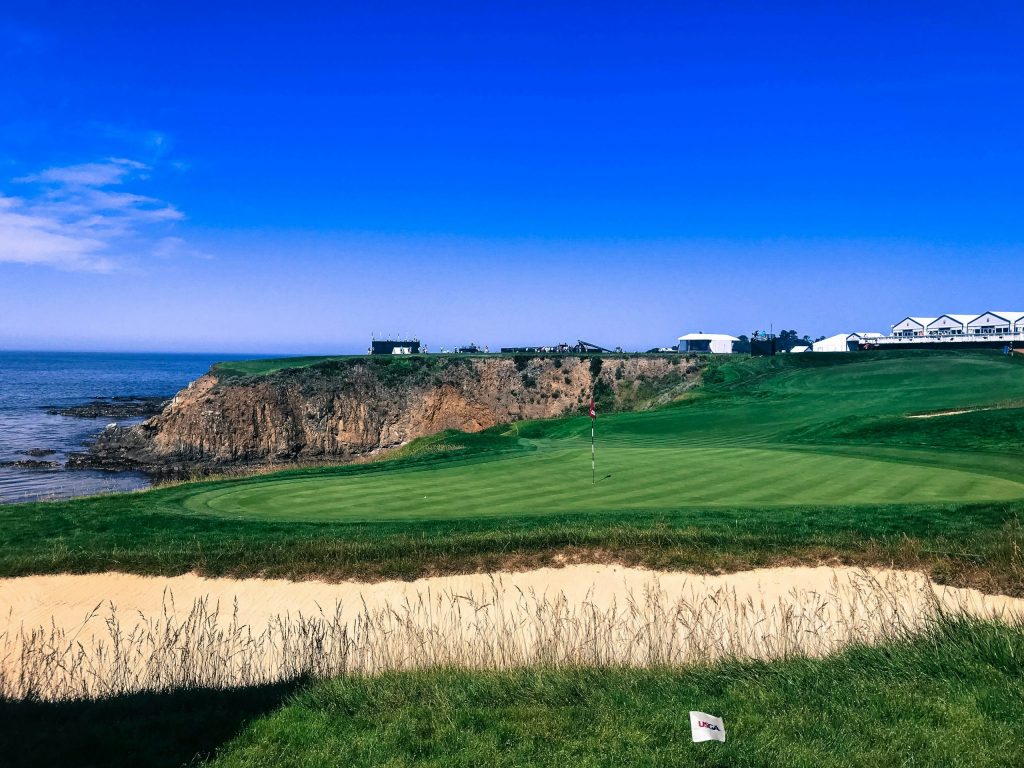 Coastal golf course with a green near a cliffside, overlooking the ocean under a clear blue sky, featuring a USGA flag and tournament tents in the distance.