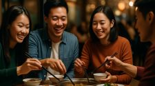 A group of four East Asian friends sit around a wooden table in a warmly lit restaurant, smiling and laughing as they share a hot pot meal with chopsticks, surrounded by various shared dishes, with the softly blurred background creating a cosy, intimate atmosphere.