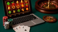 A high-resolution landscape photo showing a laptop with a slot game on screen, poker chips stacked beside it, three aces on the table, and a roulette wheel in the background.